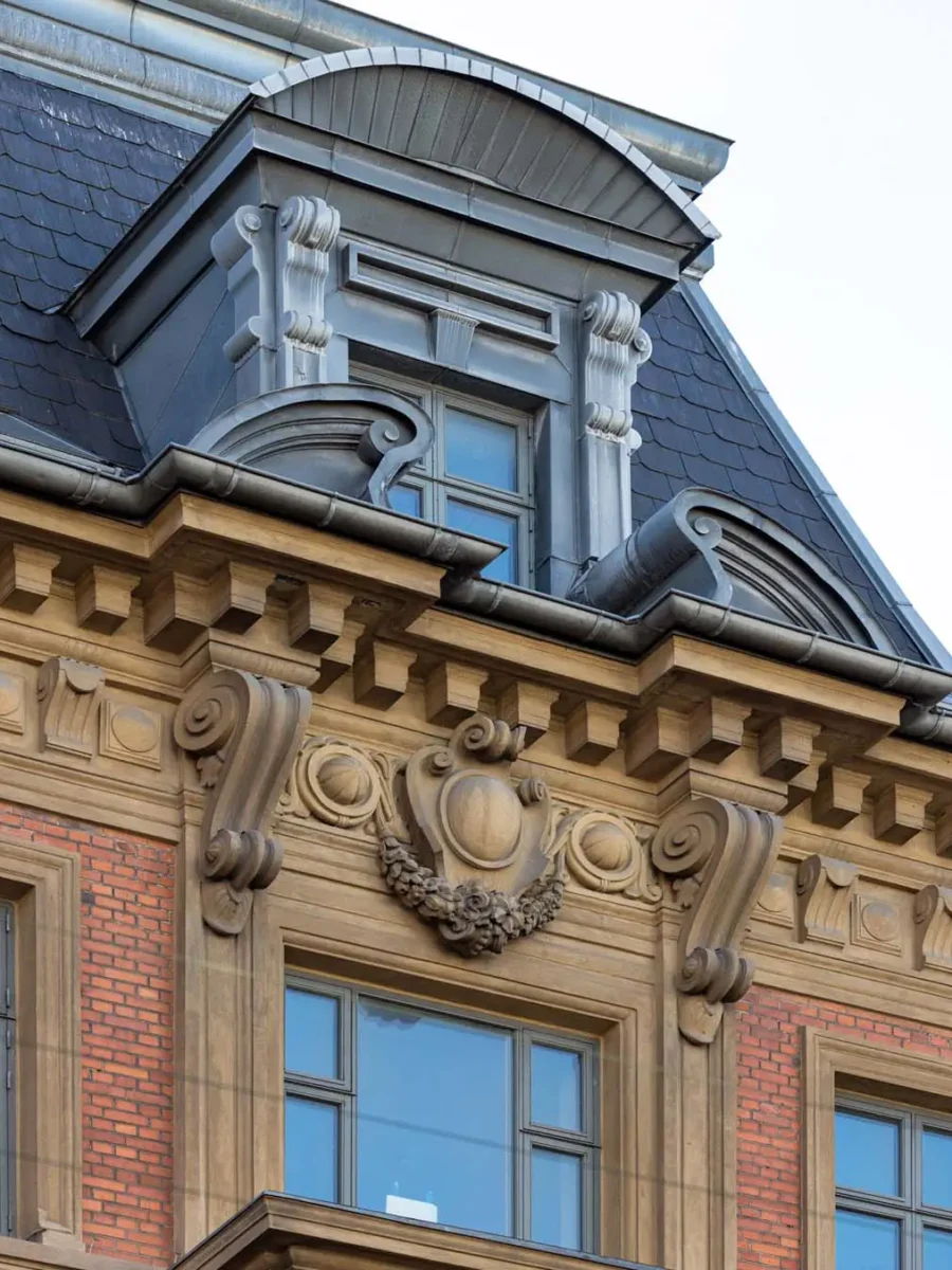 Detail einer Gründerzeitfassade: Dachgaube mit Voluten über Schieferdach, darunter Sandsteingesims mit Konsolen und einer von Girlanden gerahmten Kartusche